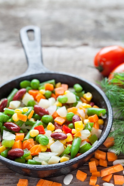 Mixed vegetable meal in old black frying pan closeup and ingredients on wooden rustic table