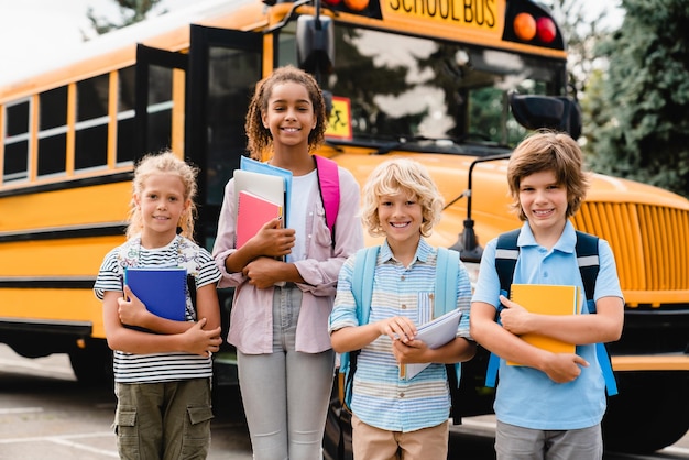 Mixedrace multiethnic school students pupils schoolchildren coming back to school after summer holidays holding notebooks copybooks next to the school bus waiting for new educational year semester