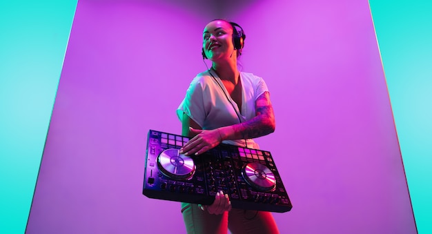 Mixing console. Young female musician in headphones performing on purple background in neon light. Concept of music, hobby, festival, entertainment, emotions. Joyful party host, DJ, portrait of artist