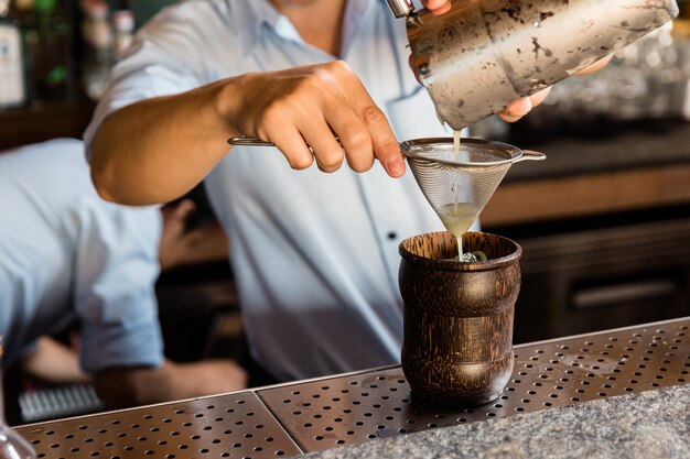 Mixoloog die cocktail met Shaker maakt, door het filter in houten drinkglas met ijsblokjes op cocktailtellerbar giet.