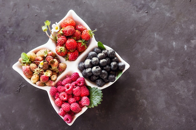 A mixture of fresh berries in a white plate on a dark gray background, natural raw nutrition