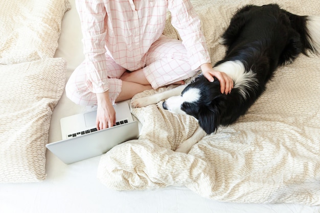 Mobiel kantoor thuis. Jonge vrouw die in pyjama's op bed met huisdierenhond werken die thuis gebruikend aan laptop PC-computer werken. Levensstijlmeisje die binnen bestuderen. Freelance bedrijfsquarantaineconcept.