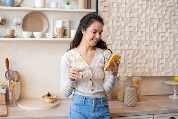 Mobiele communicatie gelukkige dame die smartphone gebruikt en zoet water drinkt in het keukeninterieur