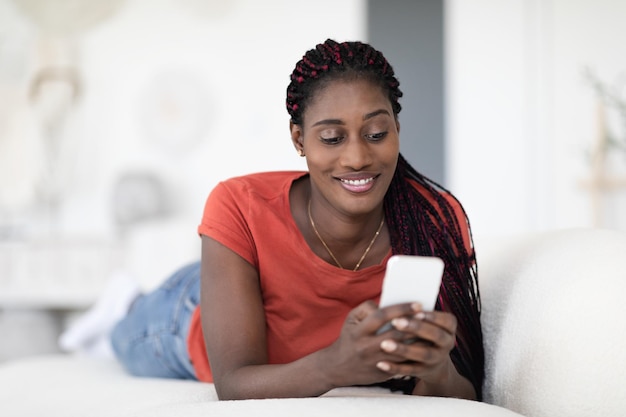 Mobiele communicatie glimlachend zwarte vrouwelijke berichten op smartphone terwijl liggend op de bank