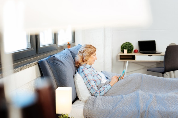 Mobiele telefoon dragen. Geconcentreerde volwassen vrouw in felblauwe pyjama die op bed leunt en nieuws zoekt op smartphone