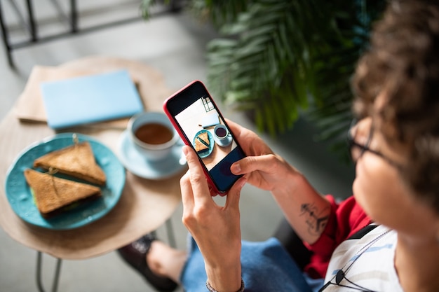 Mobiele vrouw die haar smartphone gebruikt om schot van broodjes te nemen en op lijst te drinken terwijl zij in koffie zit