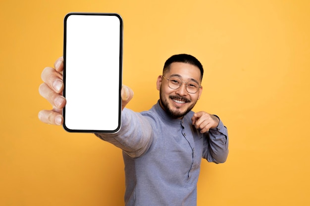 Mobile advertisement cheerful asian man pointing at big blank smartphone in hand