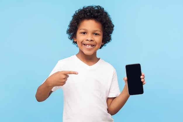 Mobile app for children. Happy excited cheerful little boy with curly hair in T-shirt pointing at cell phone and smiling at camera, showing telephone. indoor studio shot isolated on blue background