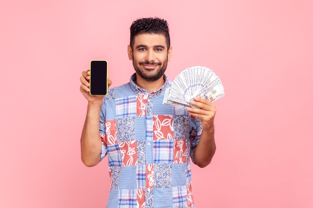 Pagamento online di servizi bancari mobili ritratto di uomo barbuto in camicia blu in stile casual con banconote in dollari e telefono cellulare con display vuoto mock up colpo in studio al coperto isolato su sfondo rosa