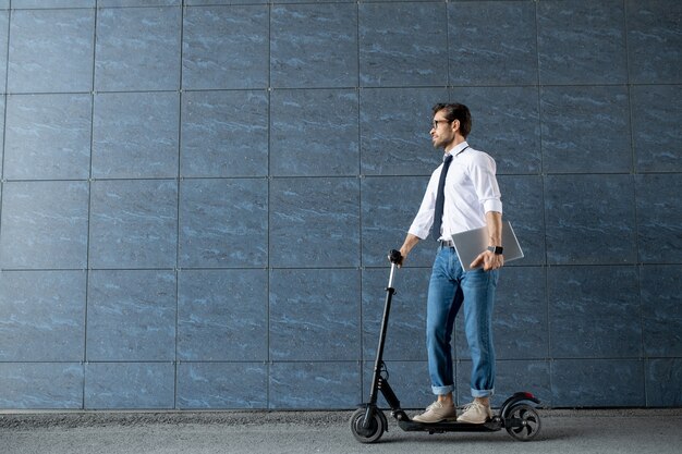 Mobile businessman with folded laptop moving on scooter along blue wall of modern building while hurrying for work