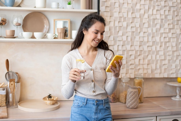 スマートフォンを使用し、キッチンのインテリアで真水を飲むモバイル通信幸せな女性