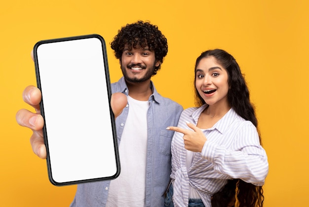 Photo mobile offer cheerful indian couple pointing at blank smartphone with white screen