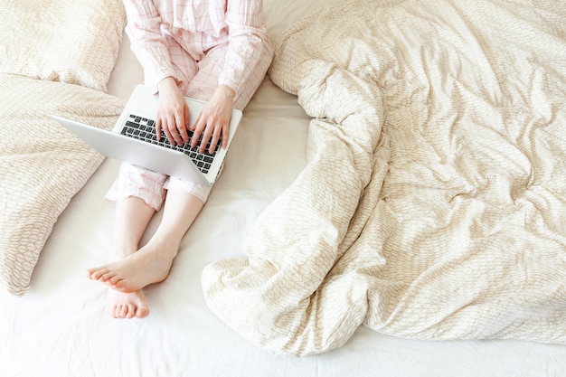 Mobile Office at home. Young woman in pajamas sitting on bed at home working using on laptop pc computer. Lifestyle girl studying indoors. Freelance business quarantine concept.