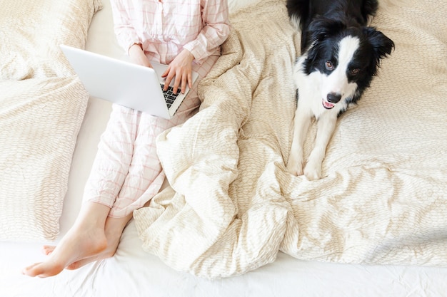 自宅のモバイルオフィス。自宅のラップトップpcコンピューターを使用して作業のペットの犬と一緒にベッドの上に座っているパジャマの若い女性。屋内で勉強しているライフスタイルの女の子。フリーランスのビジネス検疫コンセプト。