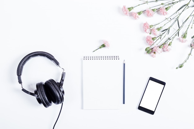 Mobile phone, black headphones, notebook, pencil and flowers lie on a white table