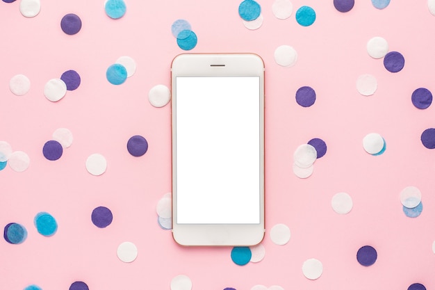 Mobile phone and blue, white and lilac confetti on pink color paper background minimal style top view