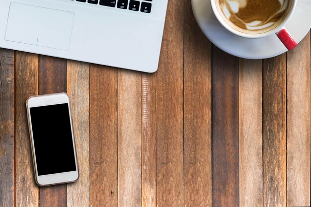 Mobile phone of empty screen and laptop and hot coffee on wooden table