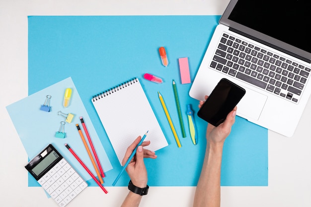 Mobile phone in hands, laptop, stationery isolated on white and blue