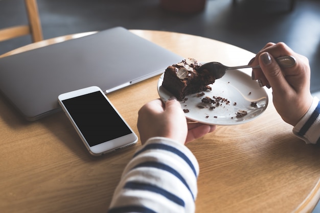 Telefono cellulare con schermo desktop nero bianco accanto al computer portatile con una donna che mangia la torta brownie