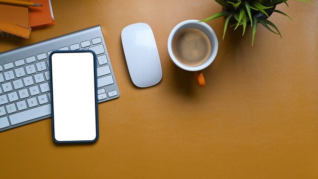 Mobile phone with blank screen, coffee cup, keyboard and plant on yellow.