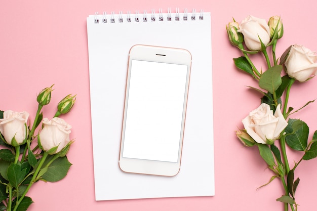 Mobile phone with a white notebook and roses flowers on pink background