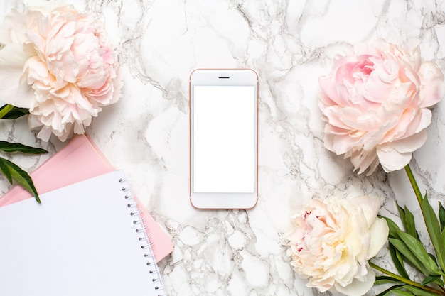 Mobile phone with a white and pink notebook and piony flowers on a marble background