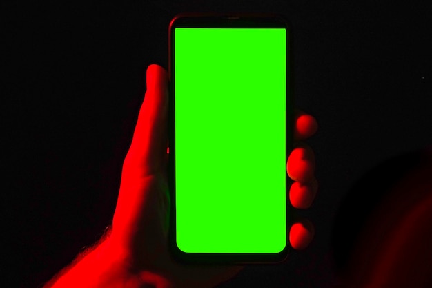 Mobile phone in young man hand Use smartphone with green screen on red blue neon lighting dark background Male showing black phone chroma key display Close up Hand lifts device up holds it still