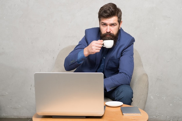 In mobile style Bearded man enjoying his hot drink and mobile internet Businessman with mobile device drinking tea or coffee at workplace Hipster in formalwear working at laptop in mobile office