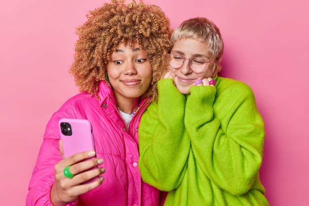 Mobile technology lifestyle and friendship concept pleased\
delighted pretty women record new content for blog via smartphone\
wear jacket and loose green jumper isolated over pink\
background