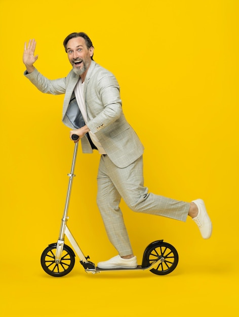 Photo mobility for middle aged business man ride scooter looking at camera and waving isolated on yellow background