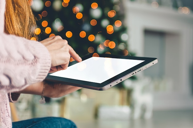 Mock up black tablet in the hands of a girl on the background, New Year Christmas tree with holiday decoration.