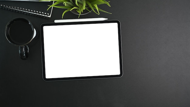 Mock up digital tablet with empty screen, coffee cup, house plant, notebook and copy space on black background.