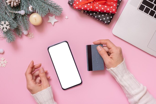 Mock up phone and credit card in female hands with Cristmas New Year work table decoration top view flat lay on pink background