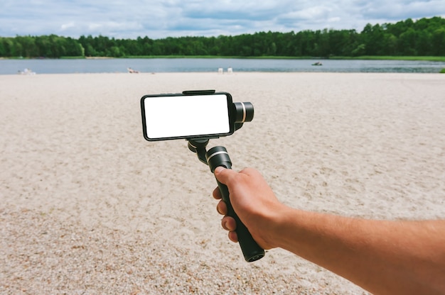 한 남자의 손에 카메라 안정기가 있는 스마트폰을 비웃습니다. 모래 해변과 호수가 있는 자연을 배경으로.