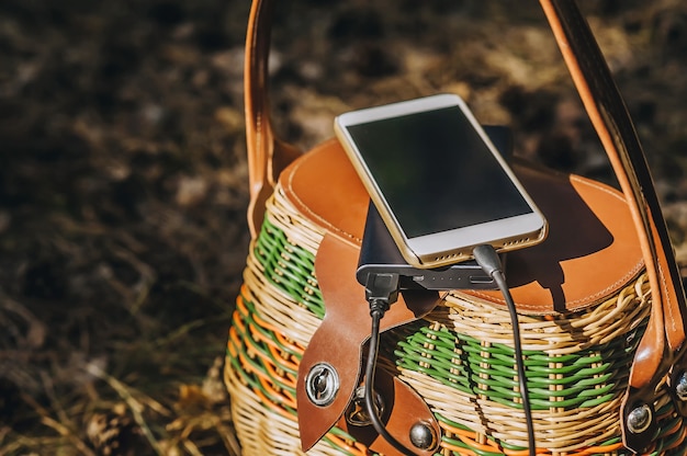 Макет смартфона с зарядкой Power Bank на корзине в лесу. Концепция на тему отдыха на природе.
