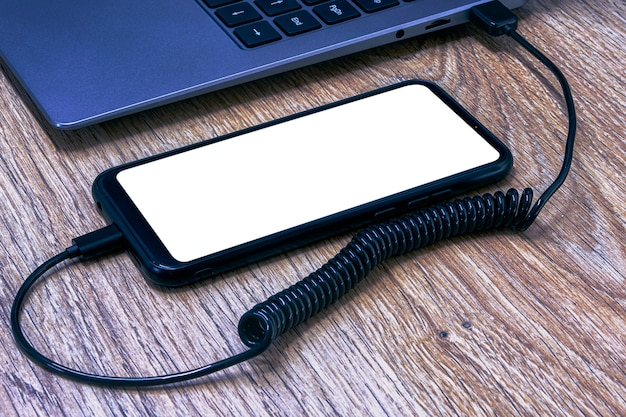 Photo mock-up of a smartphone with a white screen close-up being charged from a laptop on the background of a wooden table.