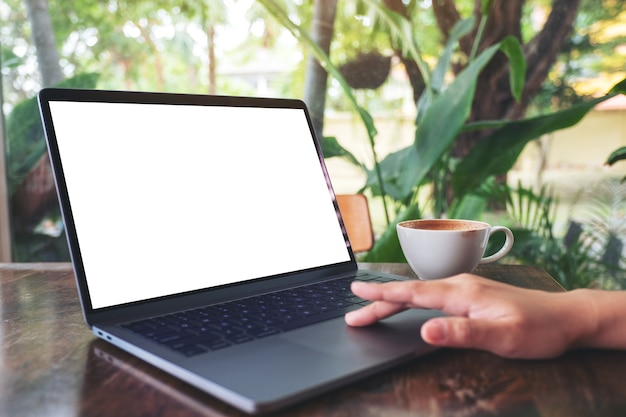 Mockup-afbeelding van de hand van een vrouw die op laptop touchpad gebruikt en aanraakt met een leeg wit desktopscherm met koffiekopje op houten tafel