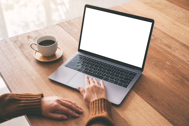 Mockup-afbeelding van de hand van een vrouw die op laptop touchpad gebruikt en aanraakt met een leeg wit desktopscherm met koffiekopje op houten tafel