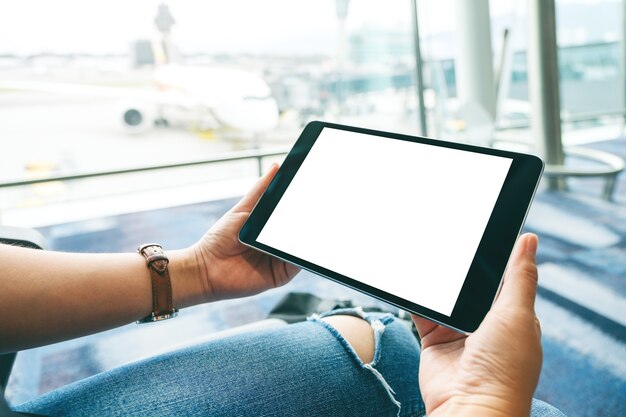 Mockup-afbeelding van de handen van een vrouw die een zwarte tablet-pc met een leeg wit scherm vasthoudt en gebruikt terwijl ze op de luchthaven zit