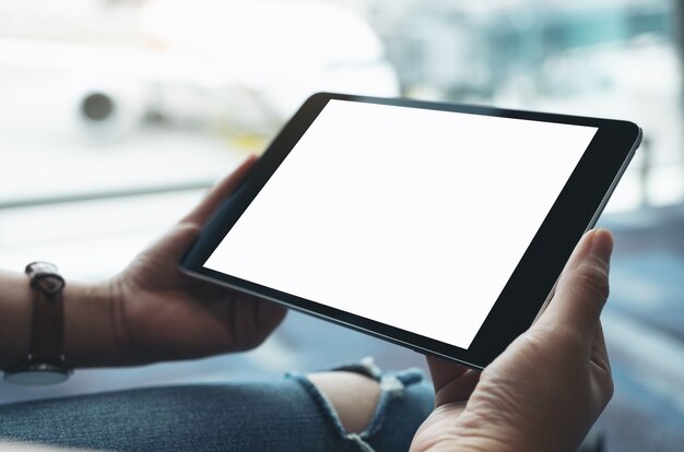 Mockup-afbeelding van de handen van een vrouw die een zwarte tablet-pc met een leeg wit scherm vasthoudt en gebruikt terwijl ze op de luchthaven zit