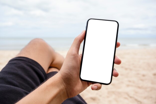 Mockup-afbeelding van een hand die een zwarte mobiele telefoon vasthoudt en toont met een leeg desktopscherm terwijl hij op de strandstoel zit