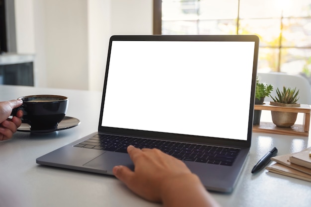 Mockup-afbeelding van een hand die het touchpad van een laptop gebruikt en aanraakt met een leeg wit bureaubladscherm terwijl je koffie drinkt