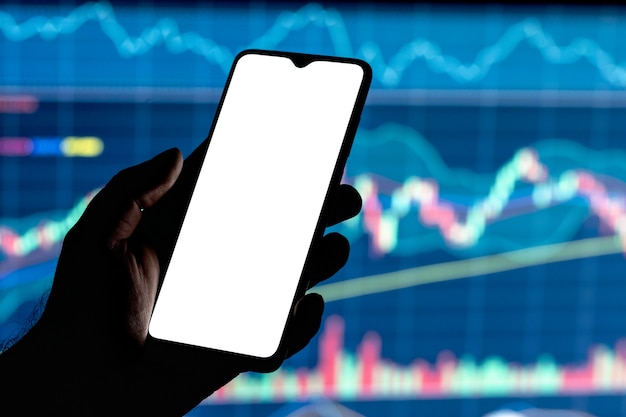 Mockup-afbeelding van een smartphone met het lege witte scherm in een hand en aandelengrafiek op de achtergrond.