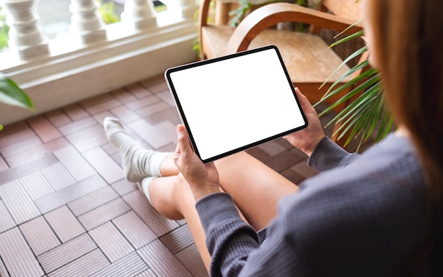 Mockup-afbeelding van een vrouw die een digitale tablet vasthoudt en gebruikt met een leeg wit bureaubladscherm terwijl ze thuis op het balkon zit