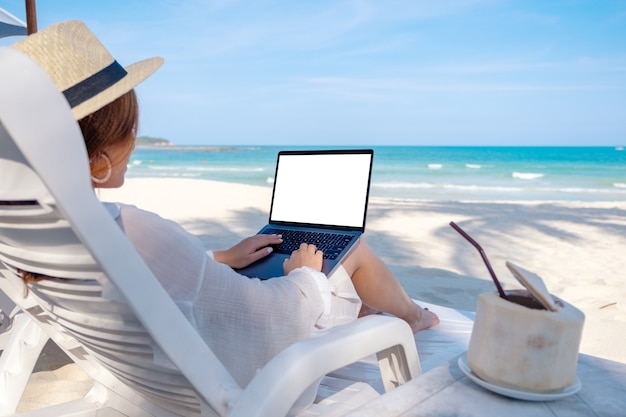 Mockup-afbeelding van een vrouw die een laptop gebruikt en typt met een leeg desktopscherm terwijl ze op een strandstoel op het strand ligt