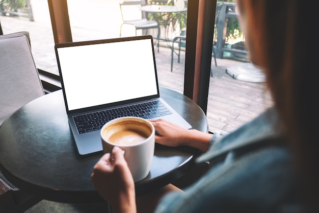 Foto mockup-afbeelding van een vrouw die een laptop gebruikt en typt met een leeg wit desktopscherm terwijl ze koffie drinkt