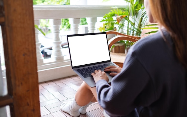 Mockup-afbeelding van een vrouw die een laptopcomputer gebruikt en eraan werkt met een leeg wit bureaubladscherm terwijl ze thuis op het balkon zit