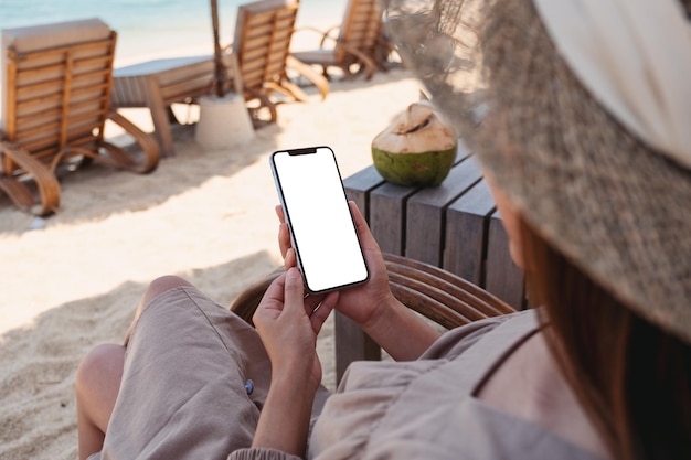 Mockup-afbeelding van een vrouw die een mobiele telefoon vasthoudt met een leeg desktopscherm terwijl ze op het strand zit