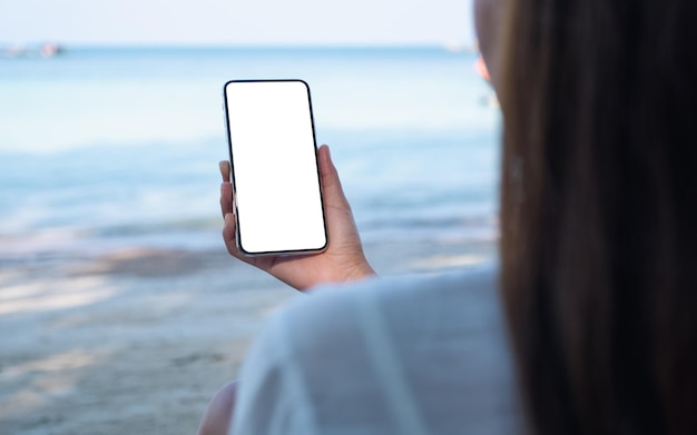 Mockup-afbeelding van een vrouw die een mobiele telefoon vasthoudt met een leeg desktopscherm terwijl ze op het strand zit