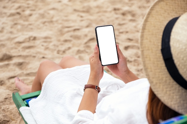 Mockup-afbeelding van een vrouw die een zwarte mobiele telefoon met een leeg desktopscherm vasthoudt terwijl ze op een strandstoel ligt